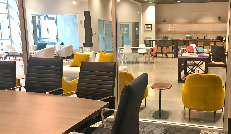 Glass-walled conference room overlooking a lounge and kitchen area with yellow accent chairs and pendant lighting.